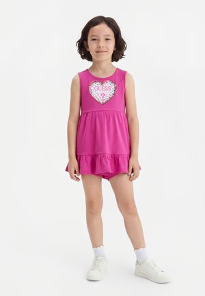 Young girl standing, wearing pink sleeveless dress with heart "GUESS" logo, white socks, and white sneakers against white background.