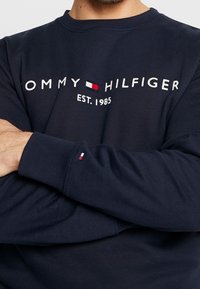 Sudadera azul marino con puños acanalados, que presenta el logo en blanco y rojo "TOMMY HILFIGER", establecido en 1985, y un pequeño detalle de bandera en la manga.