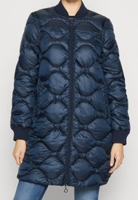 Námořnický modrý zateplený kabát s prošívaným, zaobleným designem, s předním zipem a ribovanými manžetami. Vyrobeno z lesklého, voděodolného materiálu.