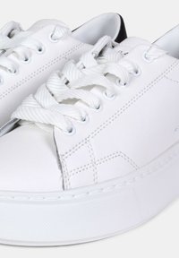 Baskets blanches en faux cuir lisse, avec un bout rond, une semelle blanche épaisse, des lacets plats en coton et de subtils détails de couture. L'arrière présente un accent noir.