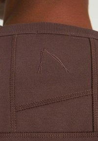 Barna textil bordázott nyakkal, geometrikus forma varrott díszítéssel és texturált varratokkal a hátoldalon.