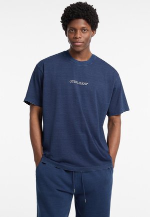 Marineblå bomulds t-shirt med rund hals, løs pasform og "GUESS JEANS" logo i hvid syning over brystet. Blød tekstur.