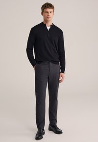 Pull noir à demi-fermeture éclair avec manches longues, associé à un pantalon gris foncé. Le modèle porte des chaussures noires et se tient avec une main dans la poche.