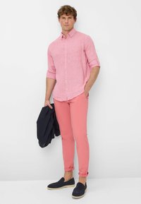 Camicia button-up rosa chiaro con taschino, maniche arrotolate; abbinata a pantaloni aderenti color corallo e scarpe slip-on blu navy, mentre tiene una giacca blu navy.