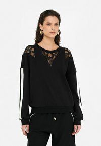 Sweatshirt noir avec un design de panneau en dentelle aux épaules et au col. Manches longues avec des rayures blanches sur les côtés, coupe décontractée.