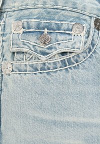 Tela de mezclilla azul claro con costuras prominentes, un pequeño bolsillo superior con botones de metal plateado y un diseño de costura distintivo.