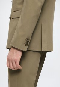 Khakifarbenes Anzugjackett mit glatter Textur, ausgestattet mit einem einzigen Schlitz, Manschette mit vier Knöpfen und tailliertem Schnitt. Sichtbare Hand, die die Seite hält.