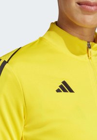Gele sportjack met een staande kraag, voorzien van zwarte accenten en een zwart geborduurd logo op de linker borst. Gestructureerde stof.