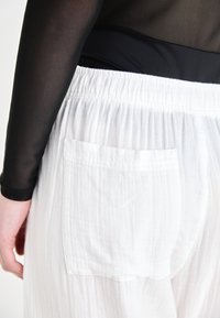 White, textured pants with an elastic waistband and a single back pocket, paired with a black long-sleeve top made of sheer material.