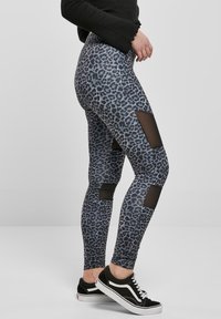 Grå leggings med sort leopardmønster, med mesh-utskjæringer på lårene og kontrasterende svarte detaljer. Brukt med svarte joggesko.
