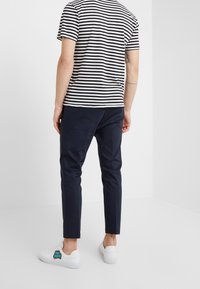 Man met zwart-wit gestreept T-shirt, marineblauwe slim-fit broek en witte sneakers met kleurrijk logo, staand in een eenvoudige witte kamer.
