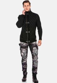 Schwarze, gestrickte Strickjacke mit hohem Kragen und Metallverschlüssen. Kombiniert mit grauen Tarncargo-Hosen und schwarzen Schnürstiefeln.