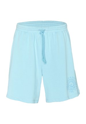Mann trägt grauen Pullover mit Ozeanwellen-Grafik und hellblauen Shorts mit passendem Logo, steht mit den Händen in den Taschen.