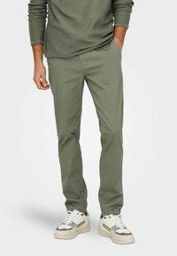 Pantalons en coton verts avec une coupe droite, dotés de poches latérales et d'une texture lisse, associés à des baskets blanches et vertes.