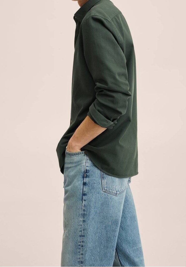 Homme portant une chemise à manches longues vert foncé avec les manches retroussées et un jean bleu clair, main dans la poche, de profil.