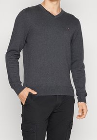 Grauer V-Ausschnitt-Pullover aus Strickstoff, mit gerippten Bündchen und Saum. Kleines Logo-Detail auf der Brust. Das Model hat die Hände in den Taschen.