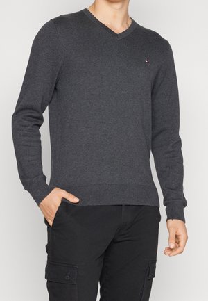 Grauer V-Ausschnitt-Pullover aus Strickstoff, mit gerippten Bündchen und Saum. Kleines Logo-Detail auf der Brust. Das Model hat die Hände in den Taschen.