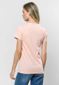 Gaiši rozā kokvilnas T-krekls ar īsām piedurknēm un apaļo kakla izgriezumu, redzams no aizmugures, kombinēts ar zilām džinsu biksēm. Gluda tekstūra.