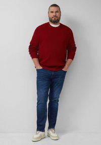 Roter Rippspullover, blaue Jeans und weiße Turnschuhe. Der Pullover hat lange Ärmel und einen Rundhalsausschnitt, während die Jeans eng an den Beinen anliegen.