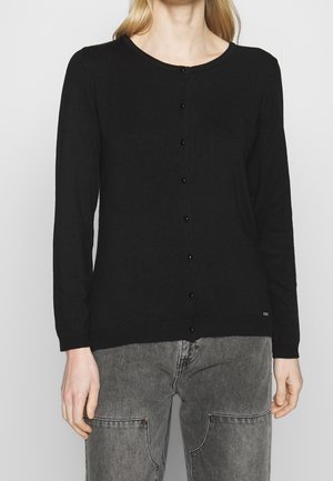 Frau trägt eine enganliegende schwarze geknöpfte Langarm-Strickjacke und ausgewaschene graue Jeans mit Fronttaschen, steht vor einem schlichten Hintergrund.