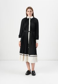 A woman stands wearing a black and white pleated dress with a belt. She has straight hair and black shoes, against a plain white background.