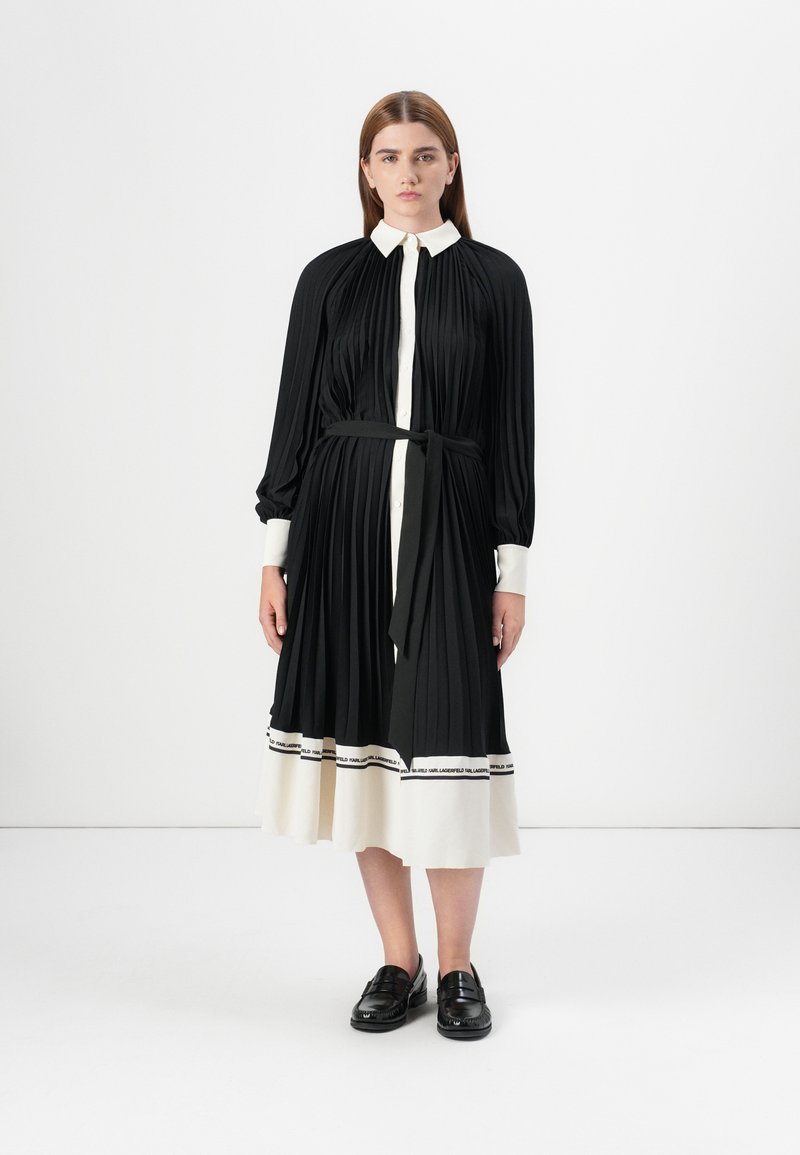 A woman stands wearing a black and white pleated dress with a belt. She has straight hair and black shoes, against a plain white background.