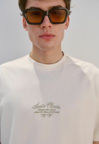 Camiseta blanca con texto bordado "Santa Clarita" en beige, combinada con gafas de sol negras y oversize con lentes marrones.
