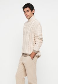 Hombre vistiendo un suéter de cuello tortuga de punto trenzado beige sobre una camisa de cuadros, con pantalones beige, de pie con las manos en los bolsillos contra un fondo blanco.