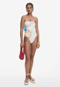 Le maillot de bain une pièce présente une base blanche avec des impressions colorées de coquillages et d'étoiles de mer. Accompagné d'un sac à main perlé rose et rouge et de sandales beiges.