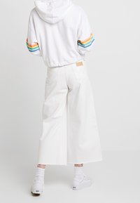 Cappuccio bianco accorciato con strisce multicolori sulle maniche, abbinato a pantaloni bianchi a gamba larga con orlo grezzo. Sneakers bianche e calzini.