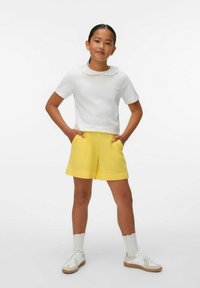 Vit kortärmad topp med spetskrage, parat med gula shorts och vita sneakers med bruna detaljer. Modellen står med händerna i fickorna.