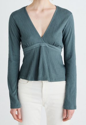 Kvinde iført langærmet, dyb V-hals tekstureret teal top kombineret med højtaljede off-white bukser, stående mod en ensfarvet baggrund.