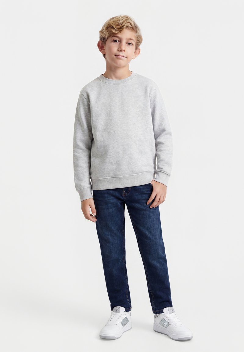 Blondais zēns stāv, valkājot gaiši pelēku džemperi, tumši zilas džinsu bikses un baltus sporta apavus, uz tukša baltā fona.