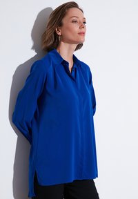 Blue, long-sleeve blouse made of smooth material. Features a classic collar and side slits at the hem for ease of movement.
