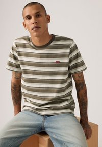 Ριγέ μπλουζάκι σε ελιά πράσινο και λευκό με στρογγυλή λαιμόκοψη, με ένα μικρό κόκκινο λογότυπο Levi's. Βαμβακερό υλικό με απαλή υφή.