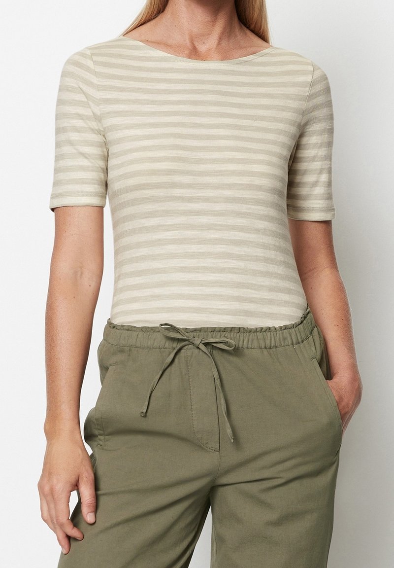 Pruhované tričko ve světle béžové a krémové barvě s kulatým výstřihem a krátkými rukávy, nošené s volnými zelenými kalhotami na šňůrku.