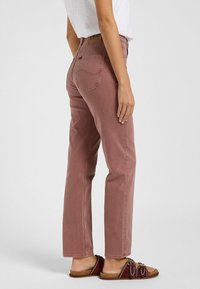 Mujer que lleva jeans rectos de color rosa polvoriento, top blanco y sandalias burdeos de tipo mocasín con hebillas, de pie contra un fondo claro y sin detalles.