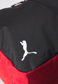 Rucsac negru și roșu cu o suprafață texturată, accente din plasă și un logo Puma alb proeminent. Design compact și funcțional.