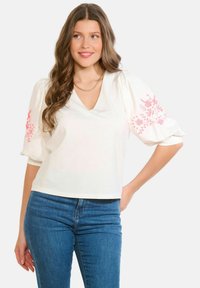 Balta blūze ar pūkainām piedurknēm un V-veida izgriezumu, piedurknēs rozā ziedu izšuvumi. Kombinēta ar zilām džinsa biksēm.
