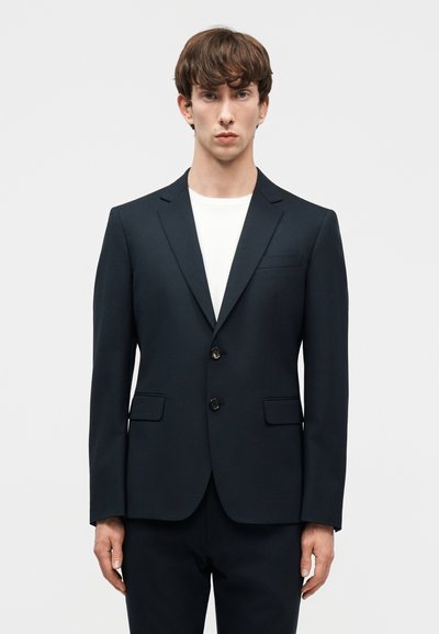 Νέος άνδρας με κοντά καστανά μαλλιά που φοράει μαύρο κομμένο κοστούμι πάνω από λευκό μπλουζάκι με στρογγυλή λαιμόκοψη, στέκεται μπροστά σε ένα απλό φωτεινό φόντο.