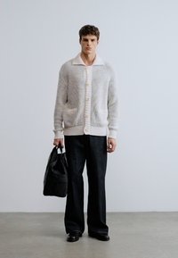 Ένα ανοιχτόχρωμο πλεκτό ζακέτα με γιακά, κλείσιμο με κουμπιά και ριμπ μανσέτες, συνδυασμένο με φαρδιά μαύρη παντελόνια και μαύρη τσάντα.