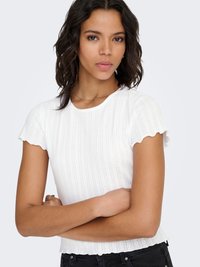 Bijeli pamučni top kratkih rukava s rebrastim uzorkom i ivicama u obliku valova, sa teksturiranim uzorkom i prilagođenim krojem. Nošen s crnim trapericama visokog struka.