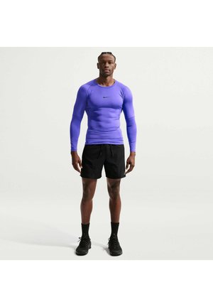 Mann steht, trägt ein eng anliegendes langärmeliges lila Sportshirt, schwarze Shorts, schwarze Socken und schwarze Sneakers vor einem einfarbigen Hintergrund.