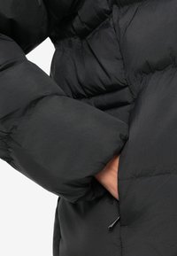 Ruka osoby čiastočne zasunutá do vrecka čiernej prešívanej bundy s vyšívaným vzorom a sťahovacou šnúrkou.