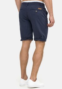 Shorts en coton bleu marine avec revers roulés, dotés de poches arrière et d'un patch logo. Le tissu a une texture lisse et un design décontracté.