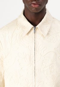 Beige broderad jacka med dragkedja, krage och intrikata bladmönster. Tyget har en texturerad, mjuk finish.