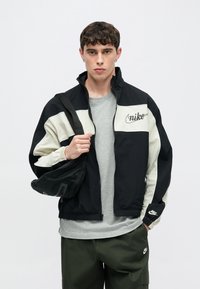 Αντιανεμικό μπουφάν Nike σε μαύρο και κρεμ χρώμα με φερμουάρ μπροστά, ελαστικά μανσέτες και διακριτικά λογότυπα. Φορεμένο πάνω από γκρι μπλουζάκι, σε συνδυασμό με παντελόνι σε ελιάς χρώμα.