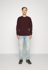 Pull en tricot bordeaux avec col rond, associé à un jean déchiré bleu clair. Des chaussures de sport noires complètent l'ensemble.