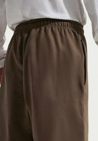 Pantalon de jogging marron avec une taille élastique, doté d'une poche zippée sur le côté et d'un tissu lisse et texturé. Chemise blanche à manches longues visible au-dessus.