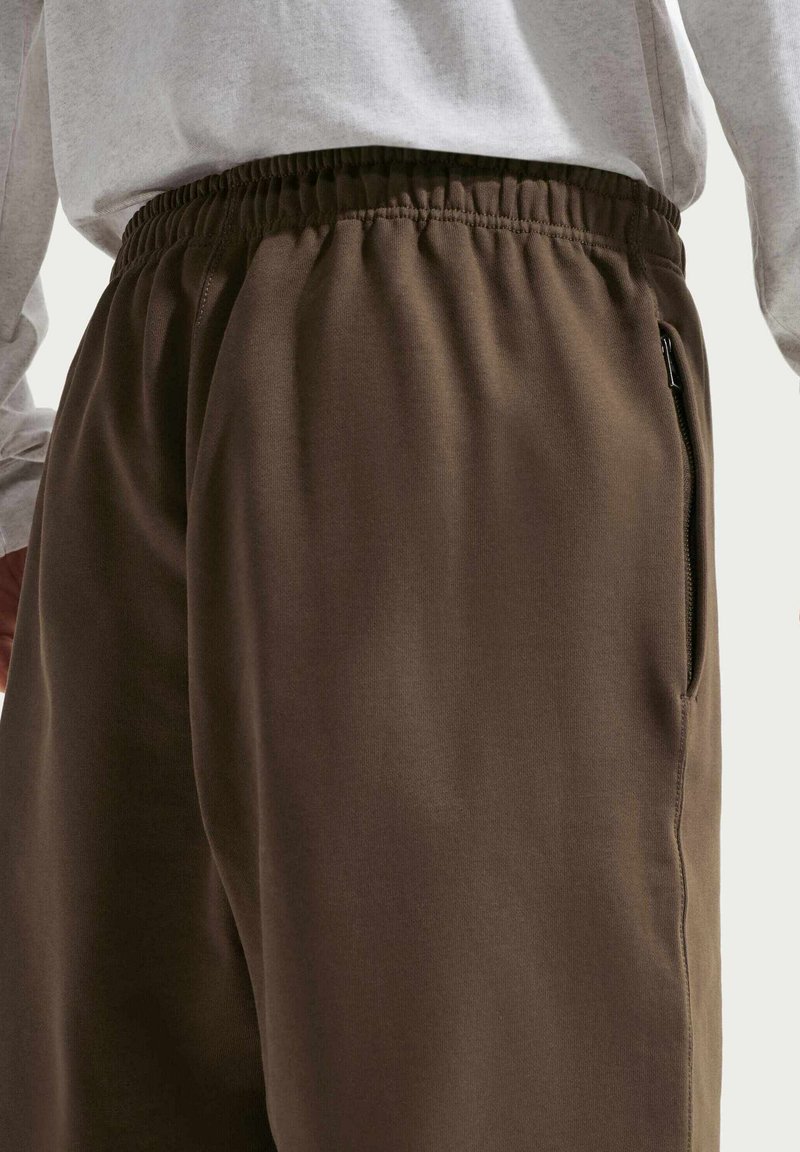 Pantaloni della tuta marroni con vita elastica, dotati di una tasca laterale con cerniera e un tessuto liscio e strutturato. Maglia bianca a maniche lunghe visibile sopra.